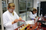 Zwei Personen in weißen Kitteln und mit Schutzbrille stehen hinter Arbeitsplatte in Labor 